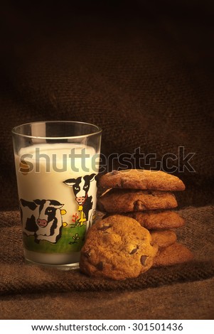some chocolate chips cookies piled, beside a small glass of milk with cows printed on it