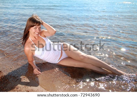A beautiful young girl dressed in white sits at the water\'s edge on the beach and smiling