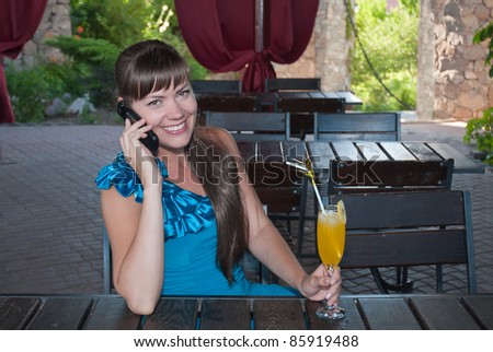 Beautiful young girl sitting at a table in a cafe and talking on a cell phone. Beautiful girl smiling. The girl in the hand of a bright yellow drink or juice.