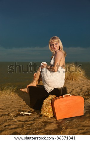 Young blonde girl in white dress sitting on a large suitcase. Next to her other bags. Against the background of dark blue clouds.