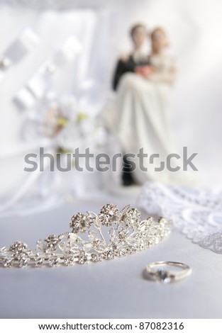Wedding silver tiara decorated with diamonds. Near the tiara ring is the bride, lace detail dress, wedding ring pillow, bride and groom figurines to wedding cake.