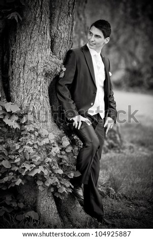 black and white photo of a handsome groom. he is leaning on a tree and looking to his bride.