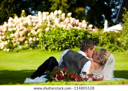 Wedding - kissing couple on meadow. Just married young wedding couple is laying down on the green meadow in fresh grass with nice colorful flowers in the background.