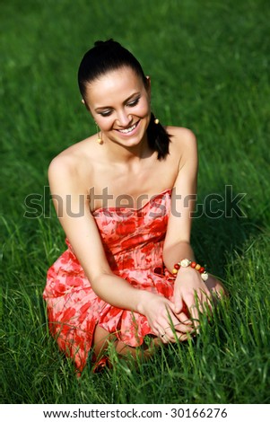 A perfect dark haired woman, wearing a nice red summer dress, is sitting on a fresh green grass meadow and relaxing in the sun