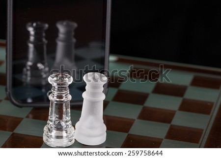 Two queens from a glass chess set on the glass board with a smart phone in front of them.  They can see their reflection in the phones screen.