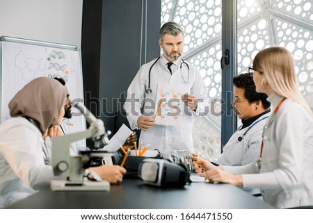 Handsome middle aged man doctor explaining information for his students at the class. Male supervisor doctor smiling while talking to his young multiethnic colleagues at the meeting