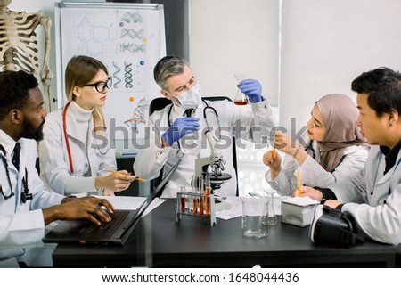 Team of multiethnic scientists in lab coats in modern laboratory conduct medical research in search of vaccine or new drug. Mature man in mask looking at flask with red liquid holding in hand