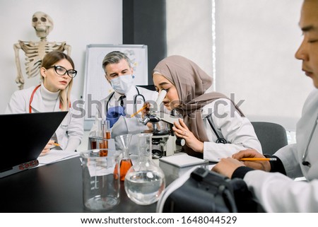 Group of scientists doctors chemists doing investigations, working in laboratory. Young Muslim woman researcher looking in microscope. her colleagues make notes