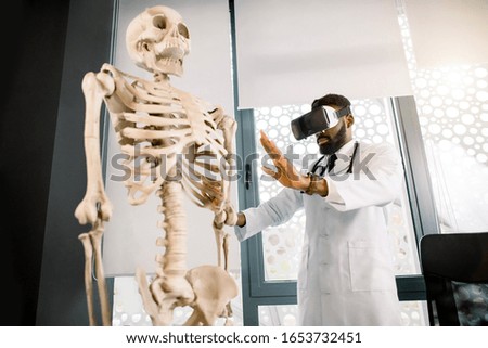 Young African man doctor, scientist or medical student uses augmented reality goggles to exam human skeleton. Virtual reality, anatomy, medicine concept