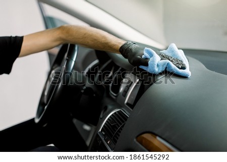 Cleaning the car, detailing concept, car wash. Close up cropped shot of hands of professional male worker, wearing rubber gloves, cleaning the interior of the car with microfiber cloth