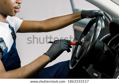 Close-up cropped image of African dark skinned worker performing professional car interior cleaning, wiping steering wheel with a brush, remowing the dust. Car service station, detailing