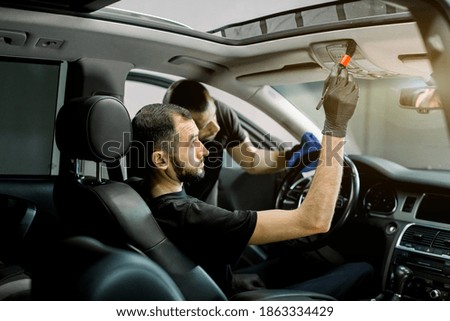 Professional car detailing service. Side view of two Caucasian men, making cleaning of modern car interior, steering wheel and dashboard with microfiber cloth and special brush
