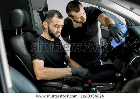 Cleaning of car interior at auto detailing service. Two professional male workers, wearing black clothes and protective gloves, cleaning control panel of the car with microfiber cloth and soft brush