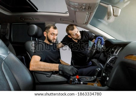 Car detailing and maintenance concept. Two male workers in black clothes, making cleaning of car interior at the car service station, one man using microfiber cloth, another one soft brush