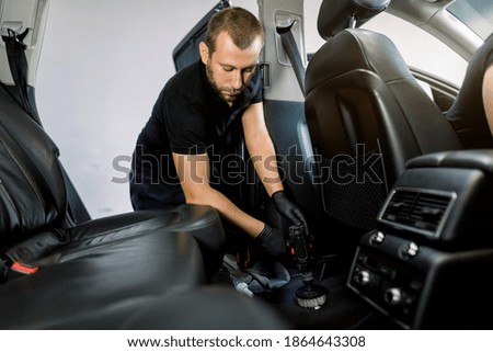 Portrait of professional young auto detailer at car service station, providing cleaning of carpet of car interior, using cleansing disinfecting foam and vacuum cleaner with brush