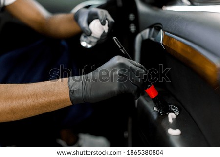Close up of hands of dark skinned car wash service worker in black protective gloves, cleaning car door with special chemical disinfecting foam and soft brush. Car wash, detailing and disinfection