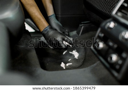 Cropped close up of hands of professional male detailer in black gloves, cleaning the carpet of car interior, applying disinfecting cleaning foam