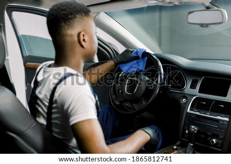 Back view of young African man worker at car detailing service, wiping car steering wheel with microfiber cloth. Cleaning car interior with sanitizer and microfiber against virus infection
