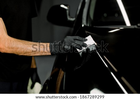 Cropped close up image of male hand in protective black glove, holding white sponge with solid carnauba wax, and polishing hood of luxury black car at professional detailing workshop. Car detailing