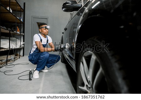 Auto detailing service, polishing of the car. Side view of young African American man worker in unifrom, t-shirt and overalls, polishing black car door with orbital polisher and wax or cream