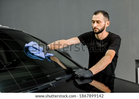 Car wash and cleaning at professional auto service station. Shot of handsome bearded young male worker in black t-shirt and gloves, cleaning car windscreen with blue microfiber cloth