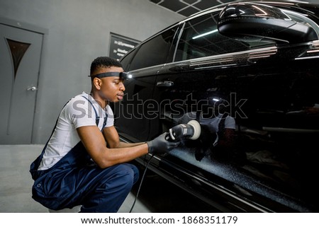 Young African man working at car detailing service, polishing black car with orbital polisher to eliminate contaminants and scratches from the surface of the car