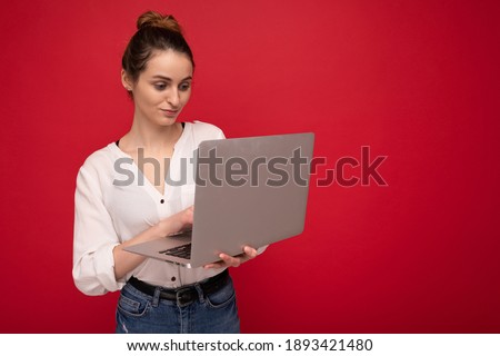 Side profile of Beautiful brunet young woman holding netbook computer looking down wearing white shirt typing on keyboard isolated on red background