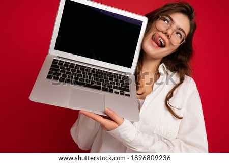 Crazy mad beautiful brunette curly young woman holding computer laptop wearing glasses white shirt showing tongue looking up to the side isolated over red wall background