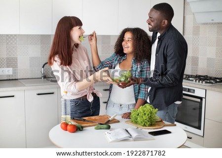 Happy teen mixed race girl feeding her mom with tasty fresh healthy food, vegetable salad