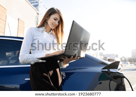 Close up side view of smiling high-skilled multitasking business lady, writing an email or reading news on laptop pc, leaning on her modern electric car at charging station