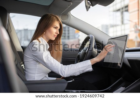 Smart electric car concept. Pretty woman in formal wear, controlling modern electric self-steering car with a digital dashboard screen, switching autopilot mode