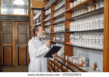 Work in pharmacy. Senior pharmacist with pen and clipboard, inspecting shelves with vintage boxes of medications, working in old ancient drugstore.