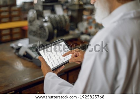 Modern and old technologies. Cropped shot of hands of senior man pharmacist working in old drugstore on a digital tablet and checking prescriptions or medicines. Vintage cash register on background.