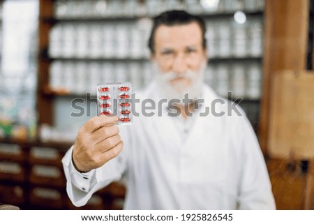 Medicine, drugs and pharmacy concept. Blurred view of friendly smiling elderly man pharmacist, demonstrating blister pack with red capsules. Old pharmacy interior.