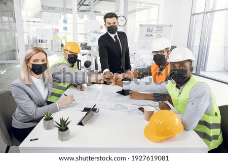Group of five multicultural people wearing black medical mask, sitting at office, bumping with fists and looking at camera. Concept of coronavirus, cooperation and construction.