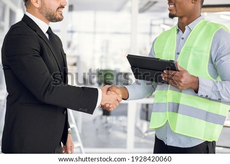 Close up of afro-american man architect handshake with his caucasian partner indoors. Two international partners using digital tablet at work. Construction industry.