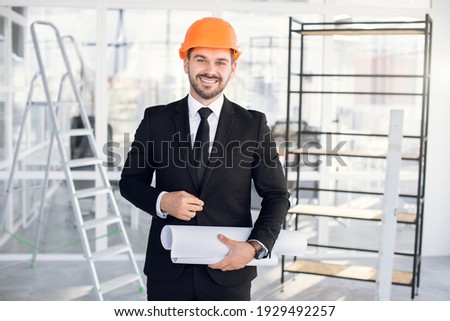 Front view of handsome man in business suit and protective helmet smiling on camera. Qualified bearded architect standing with blueprints on building object.