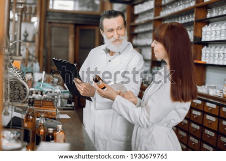 Confident female pharmacist talking with her senior male colleague about side effects of a medicine or pharmaceutical product for sale, working in old ancient pharmacy.
