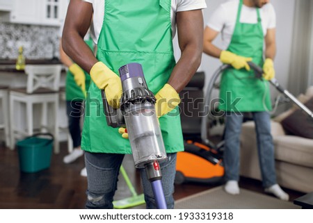 Close up of african male cleaner in uniform with vacuum cleaner in hands cleaning bright room. Team of multiracial people janitors wearing green apron and yellow gloves cleaning modern house.