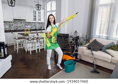 Joyful female cleaner dancing with mop during cleaning process at bright room. Pretty woman enjoying favorite work at clients house. Professional service.