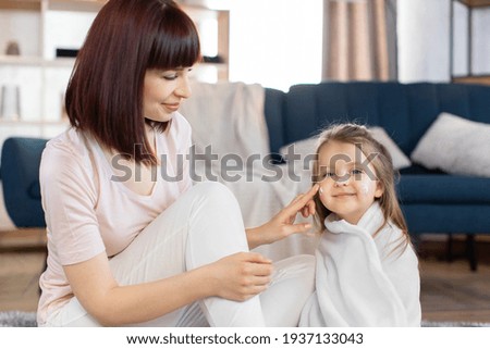 Young attractive mother taking care of her little daughter in a towel after bath, applying body lotion or cream on baby cheeks. Skin home care, children hygiene and healthy lifestyle
