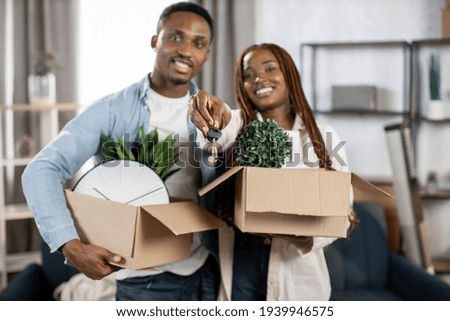 Happy african couple smiling and hugging at new apartment with boxes around. Young family showing on camera keys from flat. Concept of house ownership.