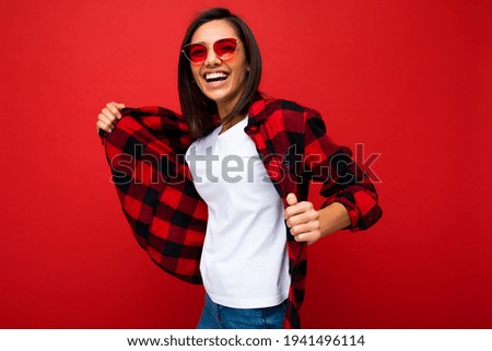 Portrait of positive cheerful smiling young brunette woman in casual white t-shirt for mockup, stylish red check shirt and trendy red sunglasses isolated on red background with copy space