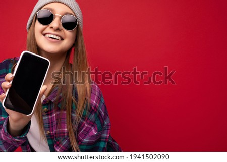 Closeup photo of attractive smiling positive young blonde woman wearing stylish purple shirt and casual white t-shirt grey hat and sunglasses isolated over red background holding in hand and showing