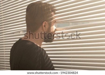 Rear view of young man with beard peeks through hole in the window blinds and looks out into the street. Surveillance and curiosity concept