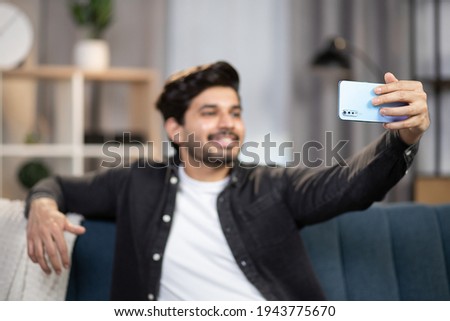 Video call, distance meeting, social networks concept. Blurred view of young Arabian man, sitting on sofa at home, having video call using smartphone, talking and having fun. Focus on phone.