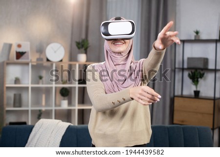 Artificial cyber world. Young excited Arabian girl in hijab and VR goggles touching air, exploring virtual reality at home, watching movie or playing interactive video game.