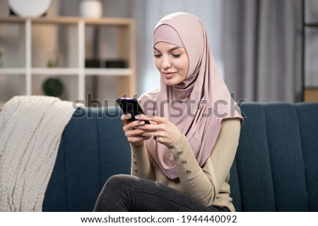 People, home and using gadgets. Pretty dreamy Arabian Muslim woman in hijab, sitting on the couch at cozy living room indoors and using smartphone for typing message or serfing social networks.