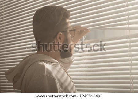 Rear view of young man with beard peeks through hole in the window blinds and looks out into the street. Surveillance and curiosity concept