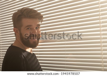 Young man with beard peeks through hole in the window blinds and looks out into the street. Surveillance and curiosity concept